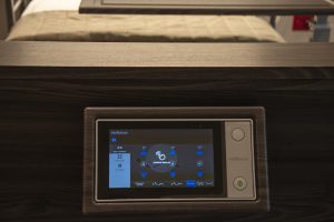 A close-up of a hospital bed's control panel. The sleek, modern panel features a touchscreen display with various icons and buttons to adjust the bed's settings, such as position and height. The bed is in the background with neatly arranged bedding.