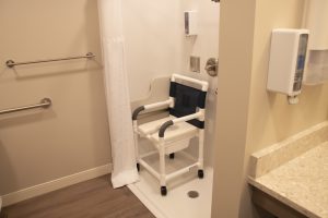 An accessible shower with a white curtain, grab bars, and a white shower chair with a blue seat and backrest. The shower has a handheld showerhead and soap dispensers. The bathroom floor is wooden, and the beige countertop has a mounted soap dispenser.