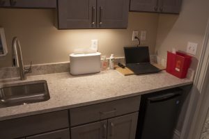 A well-lit countertop in a medical or laboratory setting, featuring a laptop, a box of consumables, cleaning supplies, and a red biohazard disposal container. The counter also has a sink, cabinets overhead, and a small refrigerator underneath.