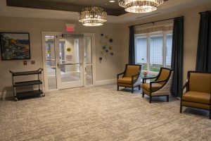 A spacious room with a neutral color scheme features two brown cushioned chairs with black frames, a small table, a wall-mounted painting, a decorative piece with colorful accents, and a rolling cart. Large windows with dark curtains allow natural light to fill the space.
