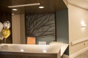 A reception desk with a beige marble countertop and an orange chair. Behind the desk is a decorative frosted glass panel featuring a branch design. To the left, a bunch of white, silver, and gold balloons are tied to the counter. Glasses are arranged on the right-hand side.