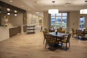 A spacious, modern dining area with wooden floors, round tables, and cushioned chairs. The room features large windows, pendant lights, and a serving counter with balloons. Hallways are visible in the background, leading to other areas.