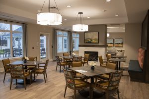 A cozy dining area with multiple square tables and cushioned chairs. Walls adorned with landscape paintings and large windows bring in natural light. Two pendant lights hang from the ceiling. Fireplace with surrounding seating area and outdoor view through windows.