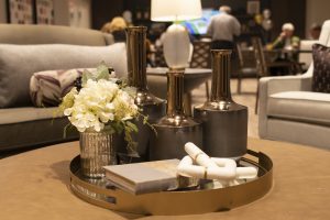 A sophisticated living room centerpiece features metallic vases, a glass of white flowers, decorative objects, and two books on a gold tray. Cozy sofas and soft lighting create a warm, inviting atmosphere in the blurred background.