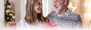 A young woman holding a gift wrapped in festive paper smiles warmly at an elderly man with a beard wearing a patterned sweater. They are standing close, sharing a joyful moment, with a decorated Christmas tree visible in the background.