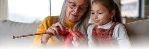 An elderly woman with long gray hair and glasses teaches a young girl with brown hair in a ponytail to knit. The woman holds a knitting needle and red yarn while the girl watches attentively. They are sitting on a couch in a cozy, bright room.