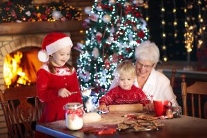 A young girl in a Santa hat and a boy in a red sweater are making Christmas cookies with their grandmother at a table. Behind them, a decorated Christmas tree and a lit fireplace create a festive atmosphere, with lights and ornaments enhancing the cozy setting.