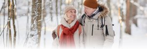 A smiling elderly couple walks arm in arm through a snowy forest. They are dressed in warm winter clothes, with the woman wearing a beige hat and scarf with a rust-colored scarf draped over her white coat, and the man wearing a brown hat and a light-colored jacket.