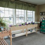 A bright room features a set of wooden plant stands adorned with various potted plants and flowers placed in front of large windows with green valances. On the right, a green shelving unit holds more plants and gardening supplies. Sunlight streams through the windows.