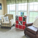 A cozy reading nook with a striped armchair on the left, a brown leather armchair on the right, and a white cubby shelf with red bins in the center. The armchairs have decorative pillows, and the shelf holds books and other items. There are large windows in the background.