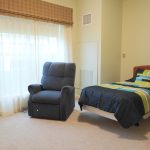 A bedroom with a single bed featuring a striped bedding set in green, blue, and white. Next to the bed is a dark blue upholstered armchair. The room has light-colored walls, a large window with sheer white curtains, and beige carpet.