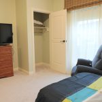 A bedroom featuring a bed with a colorful blanket, a gray armchair next to a large window with sheer curtains, a wooden dresser with a TV on top, and an open closet door showing a shelf with neatly folded towels. The room is carpeted and softly lit.