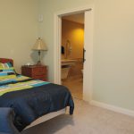 A simple bedroom with a single bed featuring a striped blue, green, and white comforter and matching pillows. There's a wooden nightstand with a lamp beside the bed. In the background, an open door leads to a dimly lit bathroom with beige tiles.