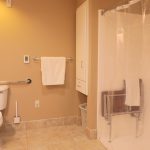 This image shows an accessible bathroom with beige walls and tiled floors. There is a toilet with a grab bar to the left, and a clear shower area with a built-in bench to the right. A towel hangs on a rack above the toilet, and a laundry basket is seen near the shower.
