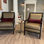 Two beige armchairs with dark wooden frames sit side by side in a room with light wood flooring. Each chair has a burgundy lumbar pillow. A small round metal table is positioned between the chairs, and a rocking chair is visible in the background near a closed door.