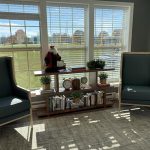 A cozy nook with two green cushioned armchairs flanking a wooden bookshelf placed in front of large, sunny windows with blinds. The bookshelf holds books, potted plants, and a Santa decoration. The view through the windows reveals a grassy outdoor scene.