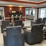 A cozy living area featuring several gray armchairs with side tables arranged in rows facing a decorated fireplace with stockings. Warm lighting and neutral tones create a welcoming atmosphere. Large windows allow natural light to enter from the right.