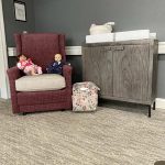 A cozy nursery corner with a maroon armchair holding two baby dolls, a floral diaper bag on the floor, and a gray changing table with a white changing pad and baby wipes on top. The room has gray walls and a light-colored carpet.
