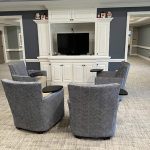 A cozy, carpeted lounge area features four gray, patterned chairs arranged in a semicircle facing a flat-screen TV mounted on a white built-in cabinet. Each chair has a small attached tray table. The room has gray walls with white trim and doors.