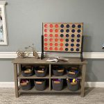 A wooden shelf against a gray wall holds a Connect Four game with red and blue discs. Below the game, the shelf has eight compartments filled with gray baskets containing various toys. A small decorative bicycle is placed on the top left of the shelf.