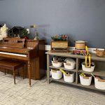 A living room scene featuring a wooden piano with a matching bench on the left and a gray wooden console table on the right. The console holds a vintage radio, a decorative jug, and several white baskets with various items on the shelves below.