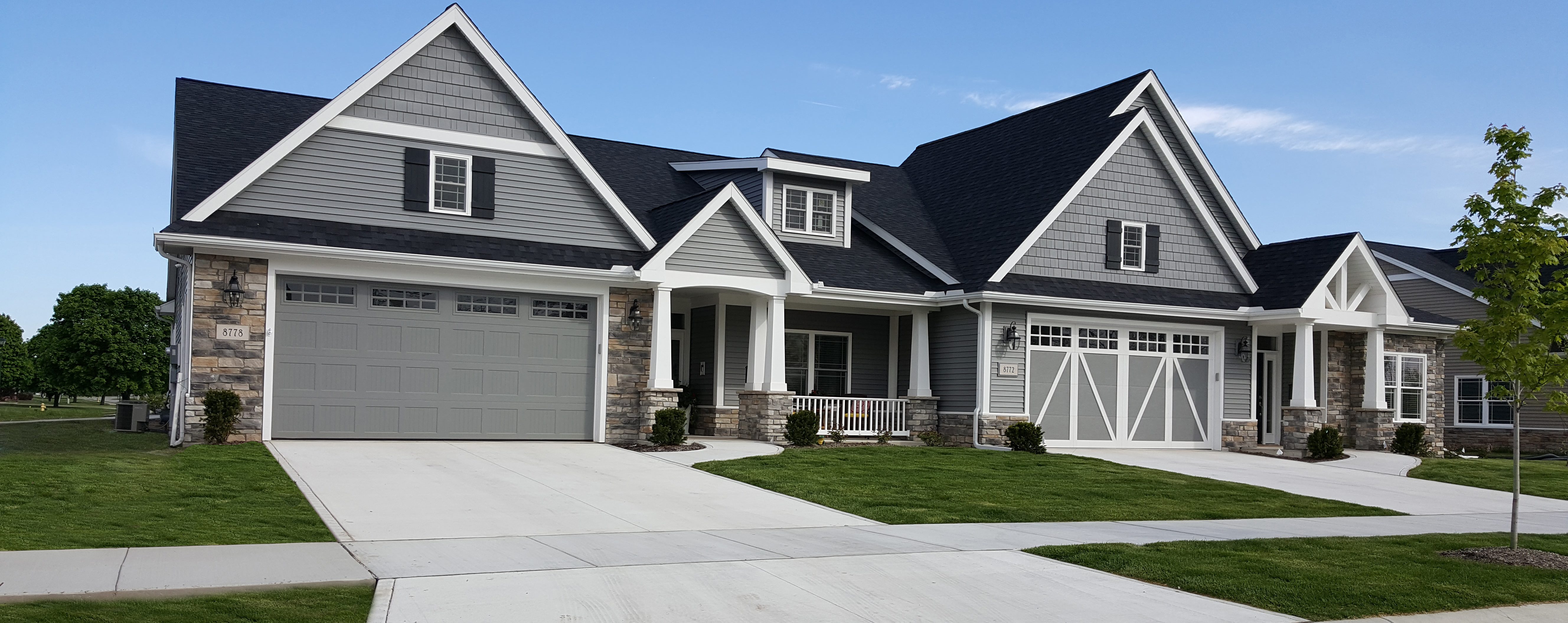 A modern suburban house with grey siding and stone accents. It has a dark shingled roof with dormer windows, two large garages with white barn-style doors, and a well-maintained lawn. A tree is seen on the right, and the sky is clear and blue.