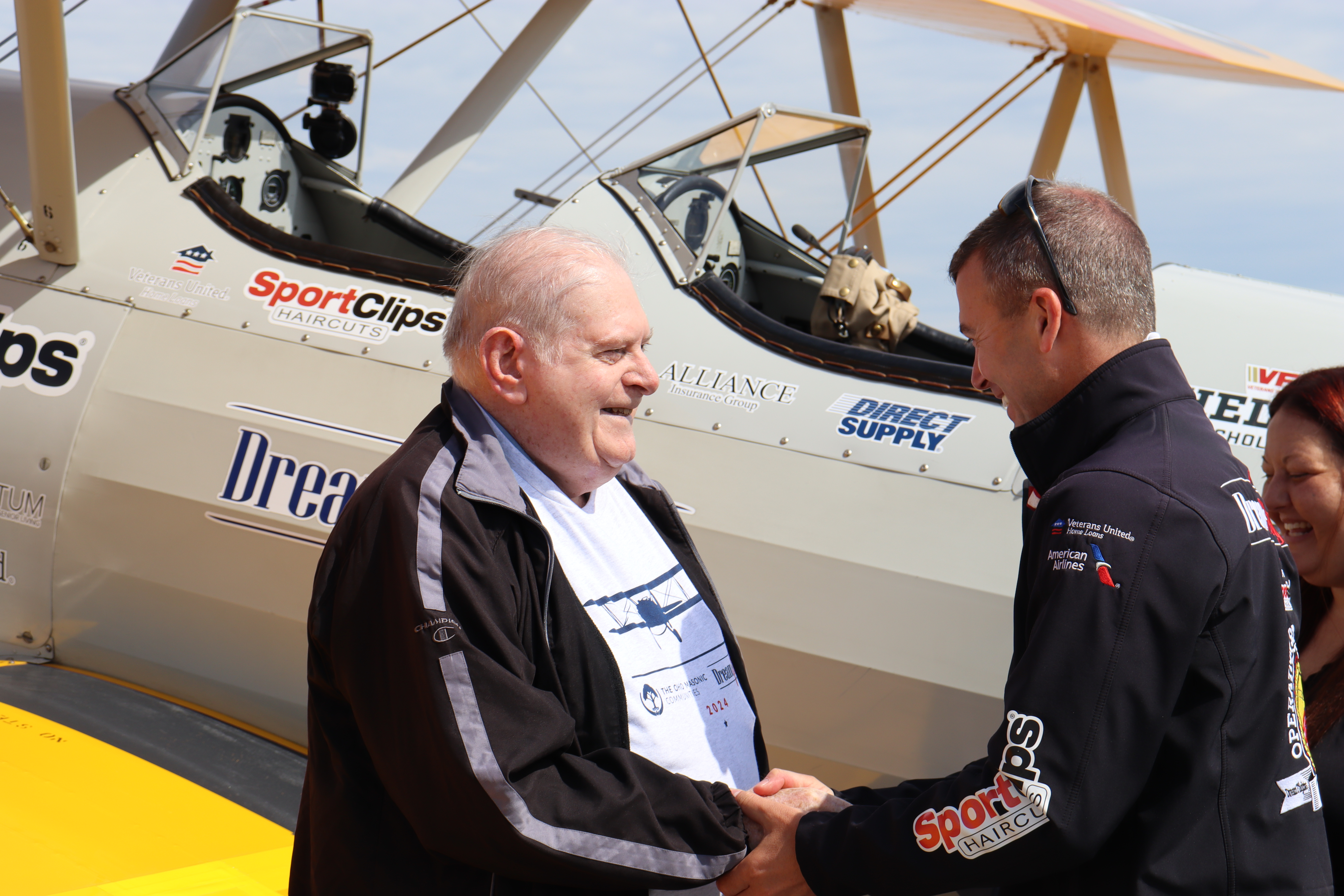  An older man wearing a black jacket shakes hands with a younger man in a dark jacket decorated with sponsor logos. They stand in front of a vintage biplane with "Sport Clips Haircuts" and "Dream" visibly marked on it, honoring veterans. A smiling woman is in the background.