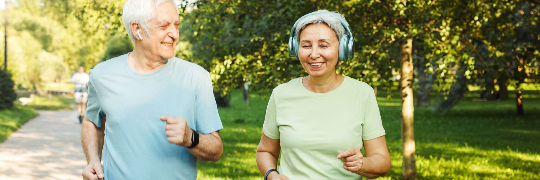 An elderly couple jogs in a park, both smiling and wearing wireless earphones. Dressed in casual athletic wear—the man in a light blue shirt and the woman in a light green shirt—they embody the vibrant lifestyle promoted by their senior living community. Trees and greenery are visible in the background.