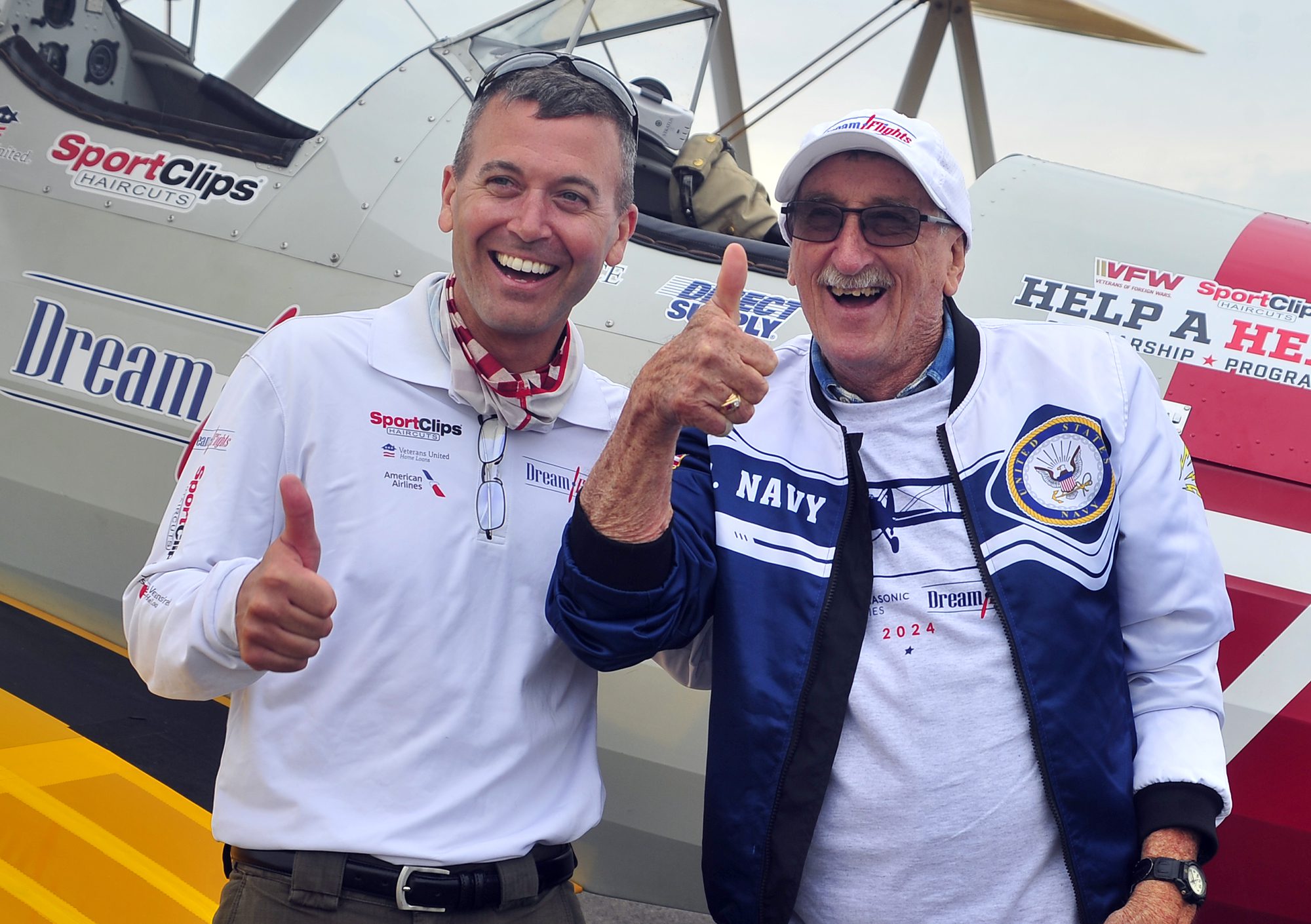 Two men are standing in front of a vintage airplane, both giving thumbs up and smiling. The man on the left is wearing a white shirt with logos, and the man on the right, dressed in a Navy jacket and cap, is honoring veterans. The airplane behind them displays several logos and text.