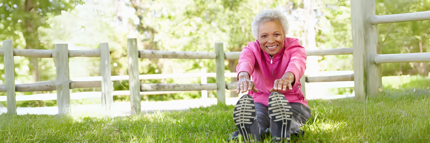  An elderly woman wearing a pink jacket and gray pants smiles while sitting on a grassy lawn at a senior living community. She stretches her legs out in front of her, reaching towards her feet. A wooden fence and green foliage are visible in the background.