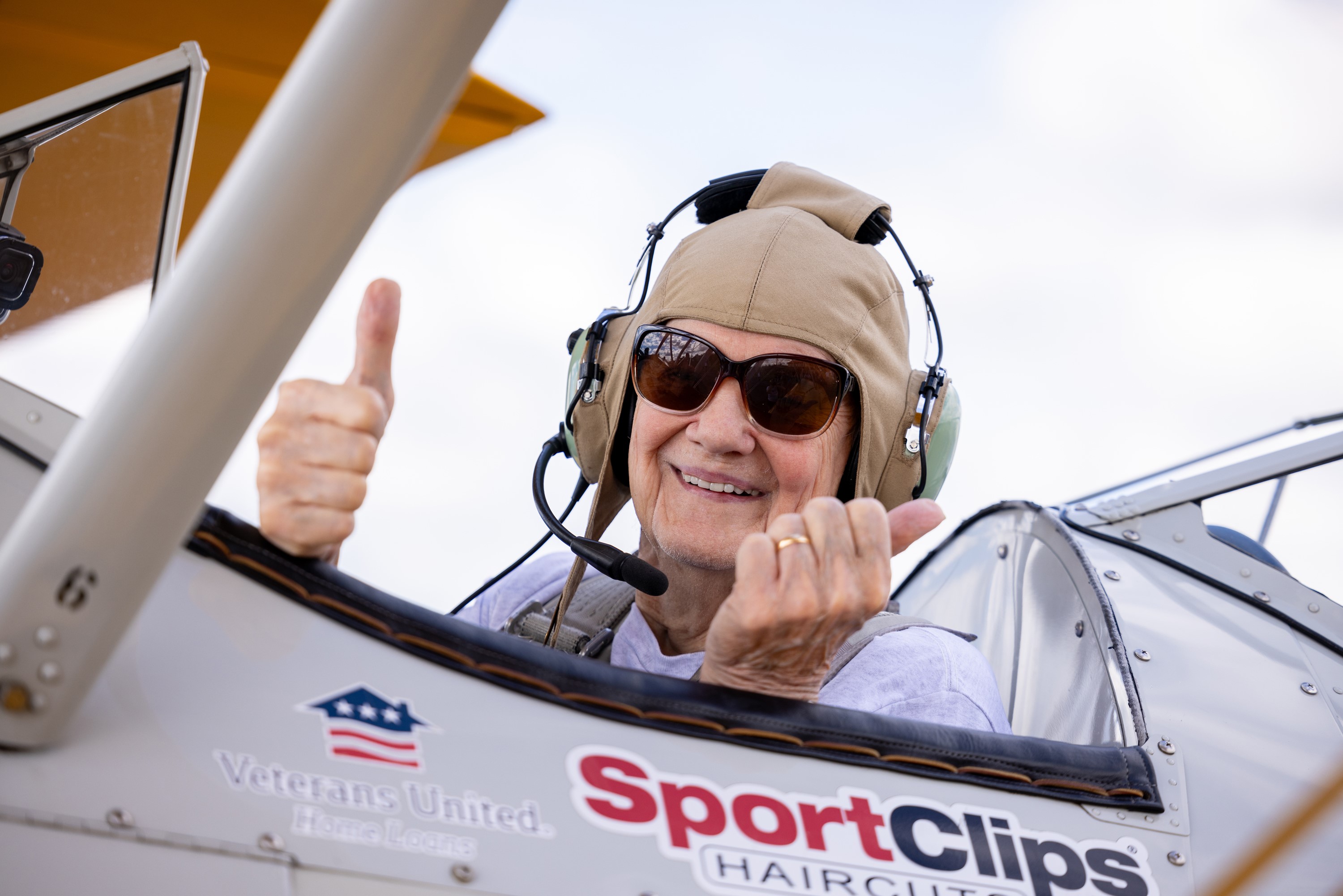 An elderly person sits in the cockpit of a vintage aircraft, giving a thumbs-up with both hands. They are wearing a flight cap, sunglasses, and a headset. The airplane is branded with "Sport Clips Haircuts" and "Veterans United Home Loans" logos, honoring veterans from the senior living community.
