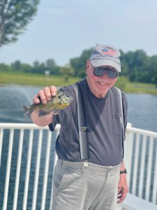 An elderly man in sunglasses, a baseball cap, and suspenders is standing on a dock in front of a private pond, holding up a small fish proudly. Trees and greenery are visible in the background under a clear sky.