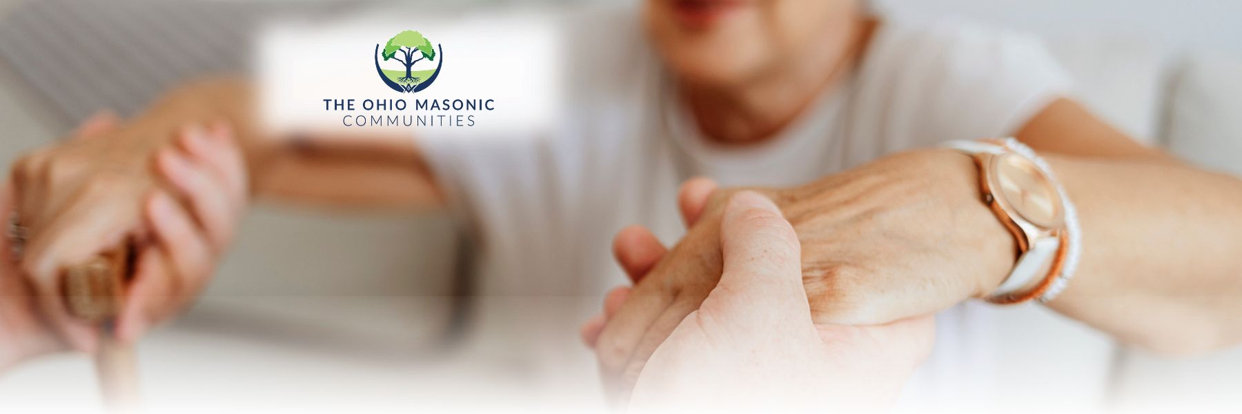 A close-up shot of an elderly person's hands being gently held by another pair of hands. The elderly person is wearing a watch. The logo for "The Ohio Masonic Communities Senior Living" is visible in the background, featuring a tree inside a circle with hand figures.
