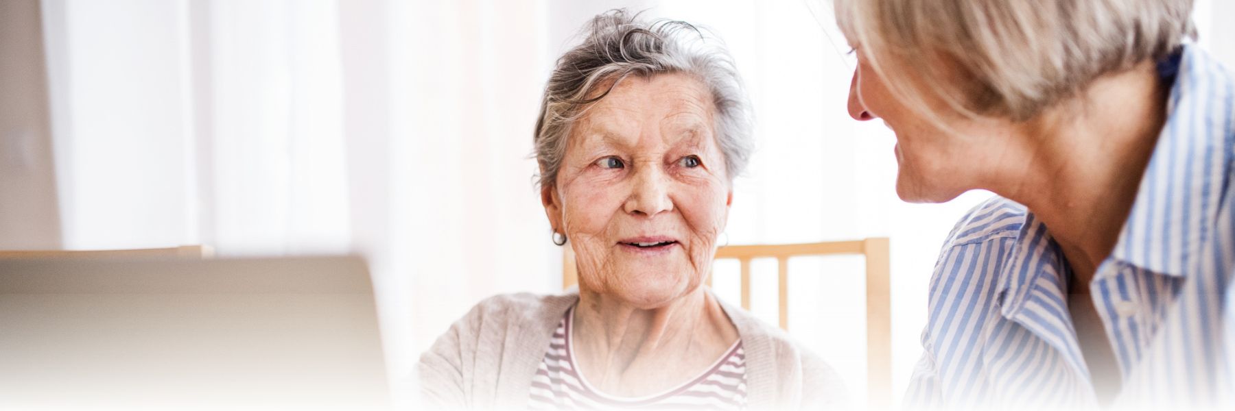  An elderly woman with gray hair, wearing a striped shirt and light sweater, smiles while conversing with a younger woman with short blonde hair in a blue striped blouse. They are seated indoors with soft, natural lighting, creating a warm and inviting atmosphere typical of senior living.