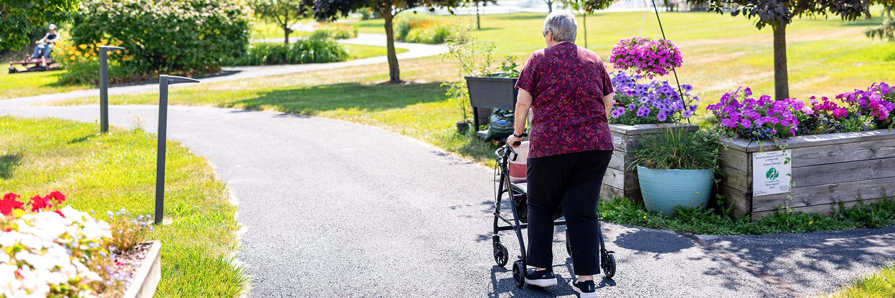 Senior woman walks with mobility device on paved road with flowers and trees on sunny day
