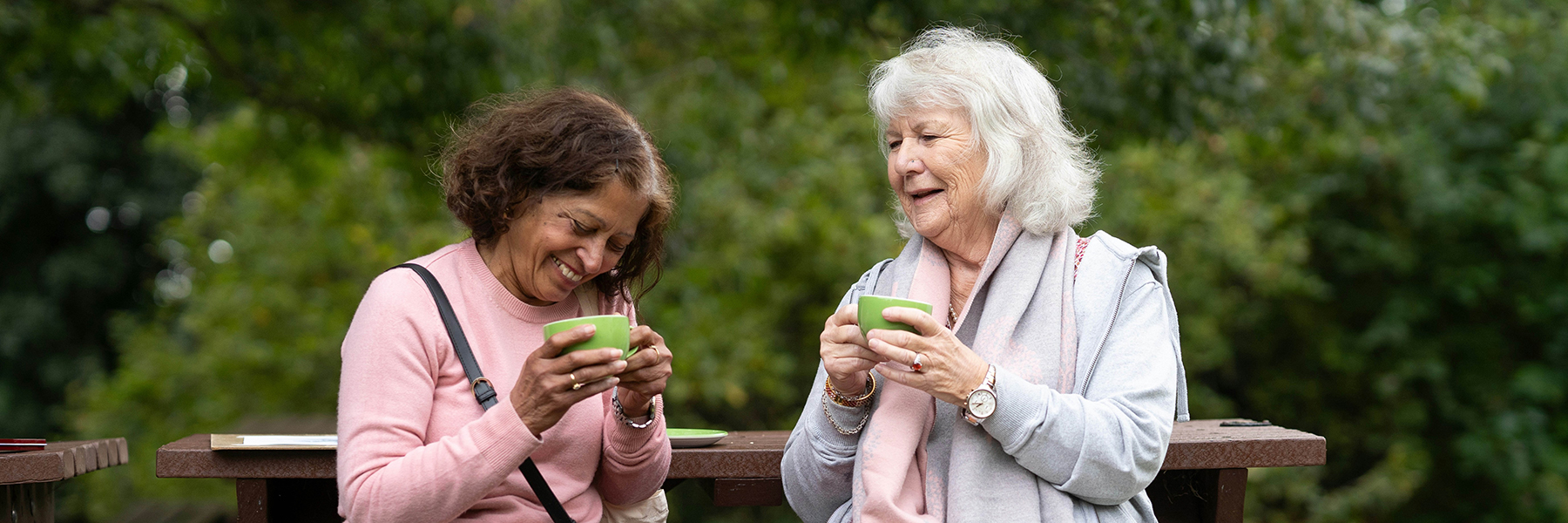 Two older adult women sit outside by greenery, laughing and holding two green mugs