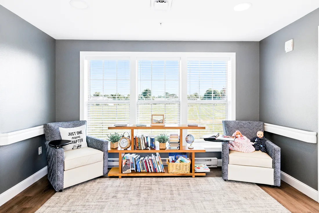 Memory support room with 2 chairs, 2 dolls, and a shelf with books