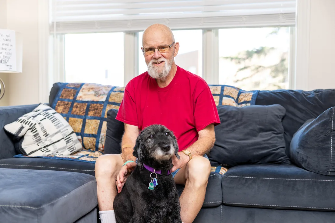Older adult male sits on a couch by a black dog