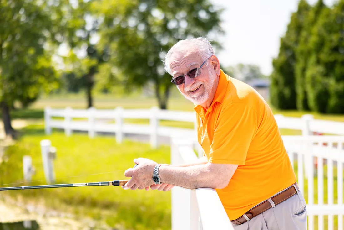 Older adult male holds a fishing pole outdoors over a white fence