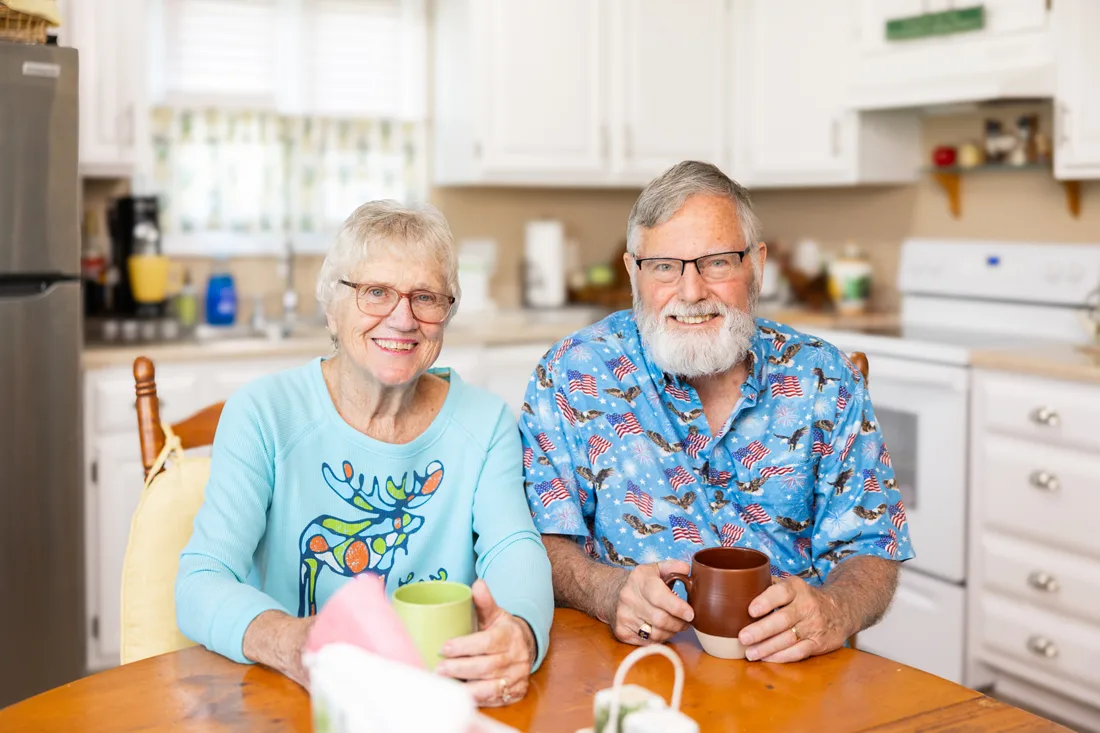Older adult couple sits in a kitchen holding coffee cups
