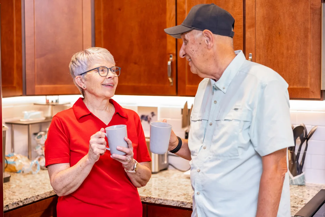 Older adult female and male chatting while holding coffee mugs in the kitchen