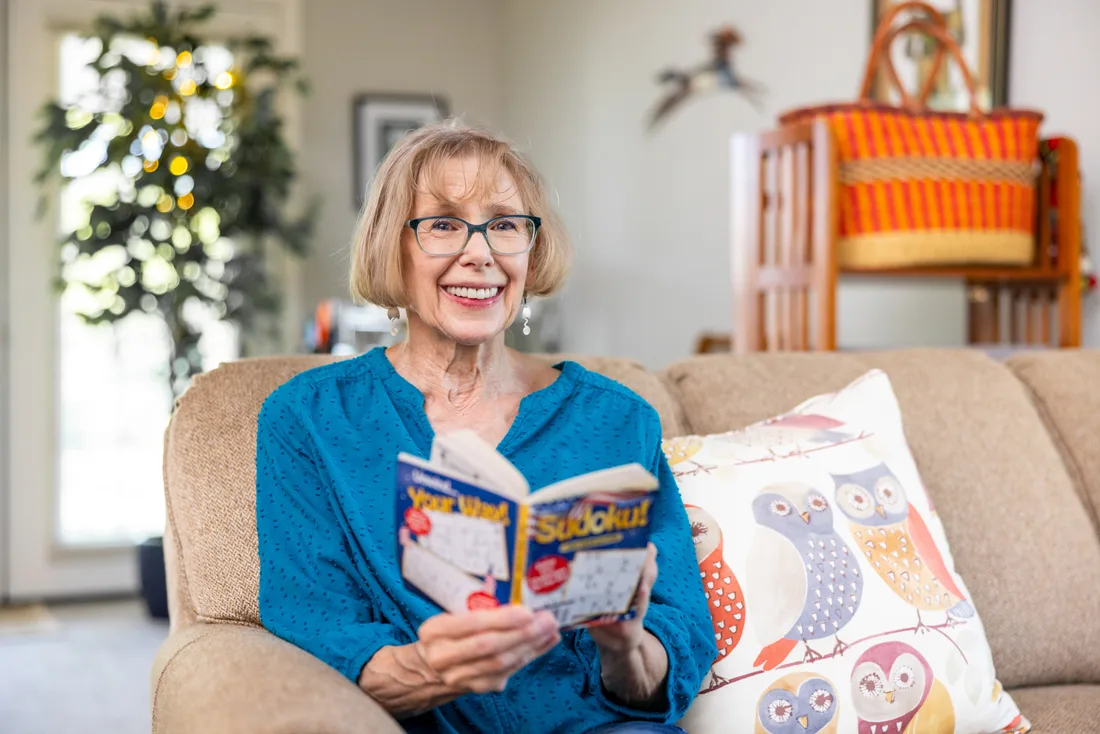 Older adult female holding a Sudoku book while seated on a couch