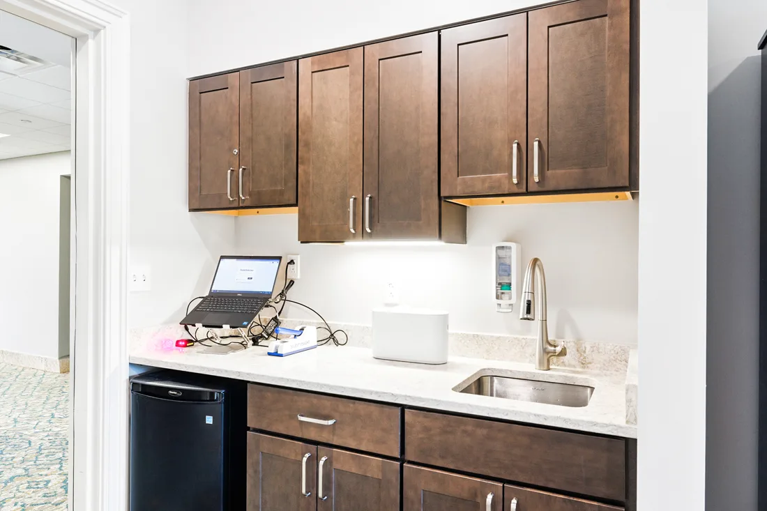 Area with cabinets, a sink, a small refrigerator, and a laptop computer