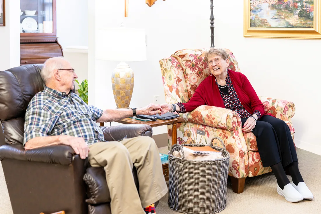 Older adult male and female sits on a chair and smiles while holding hands