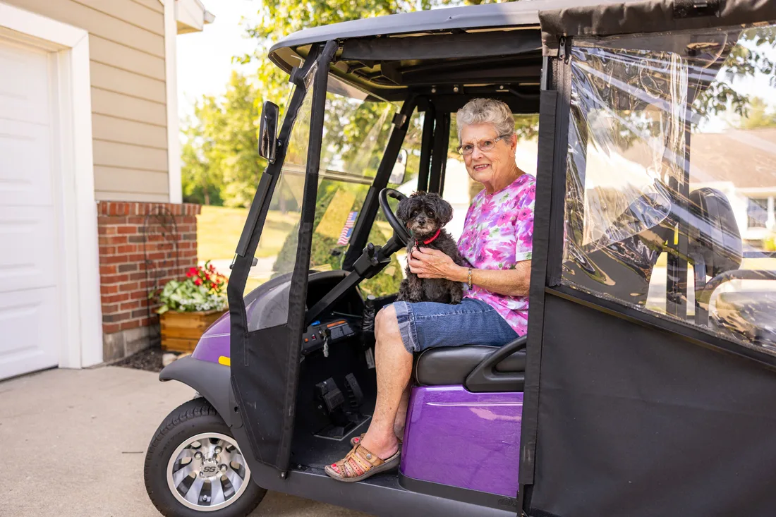 Older adult female holds a dog on her lap while sitting outdoors in a golf cart