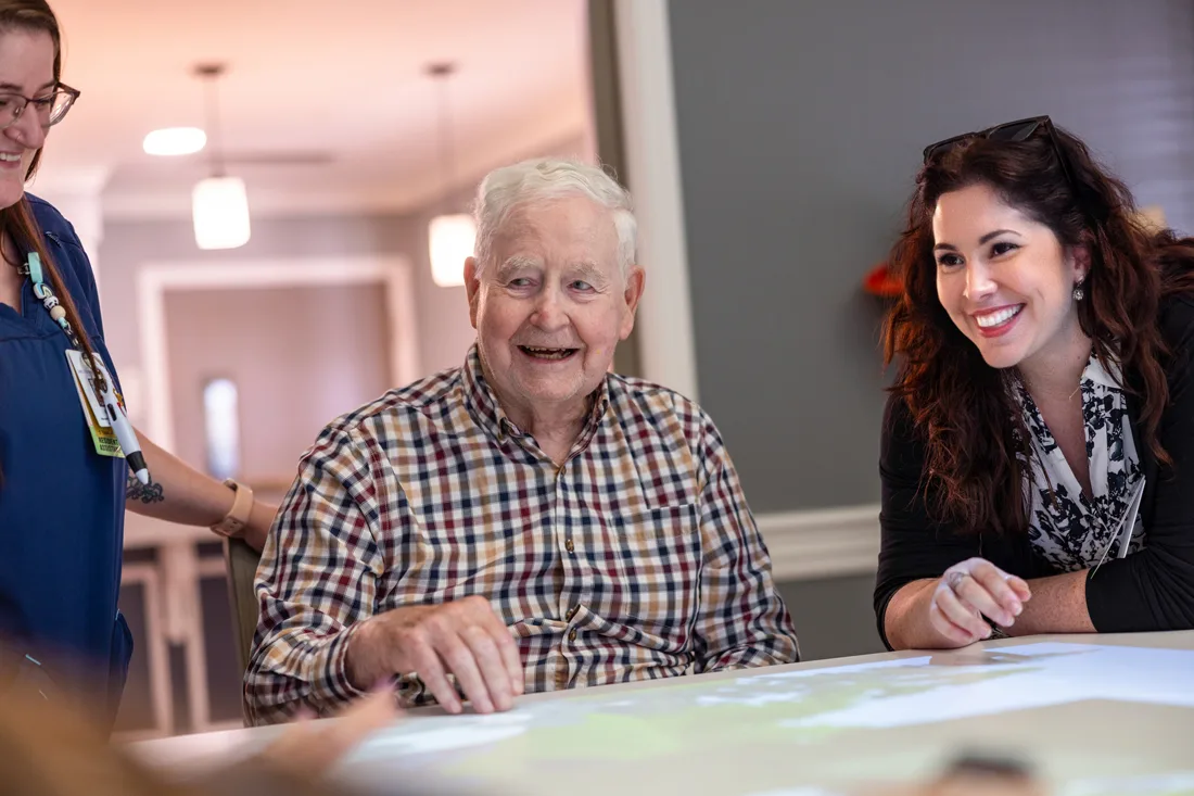 Staff members help an older adult male interact with digital images on a large table