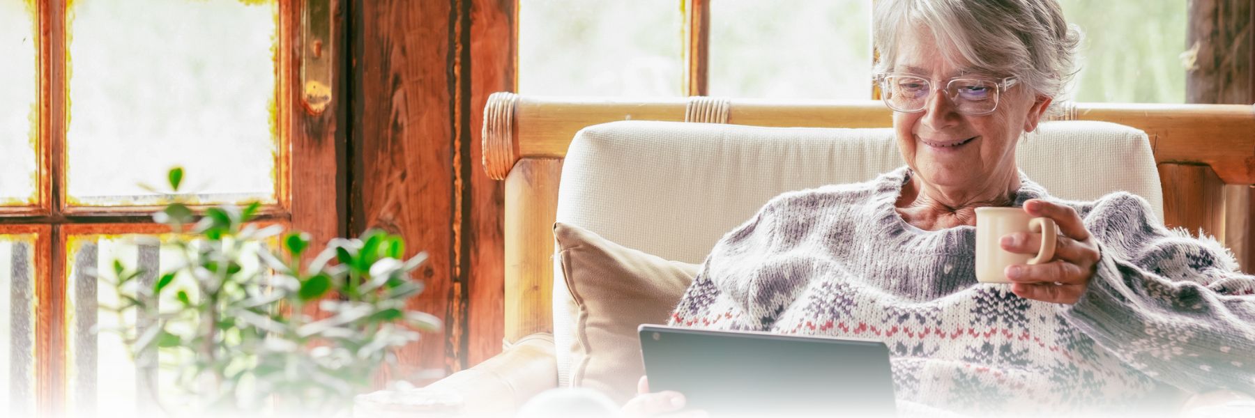 Older adult female drinks from a coffee mug and looks at a tablet by some windows