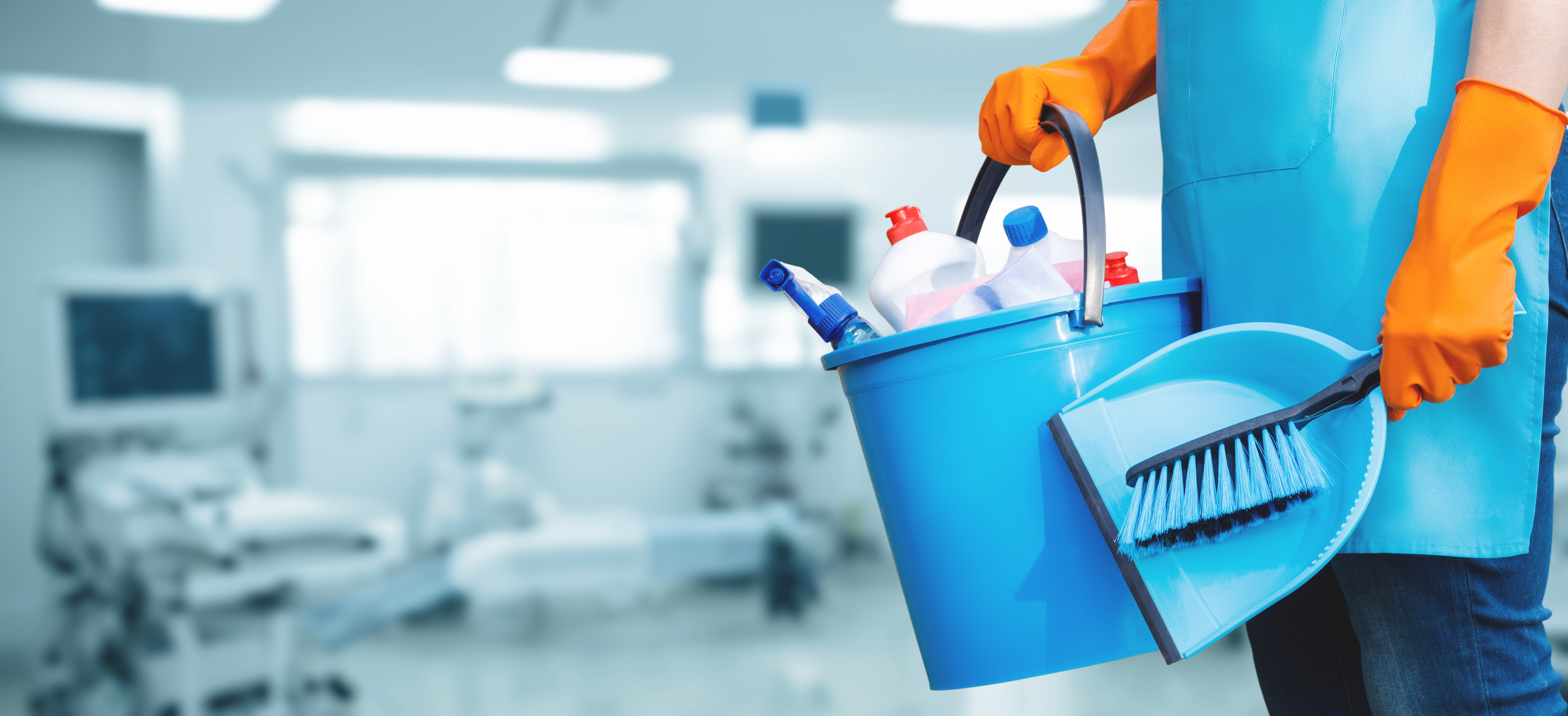A person wearing orange gloves holds a blue bucket filled with cleaning supplies and a dustpan with medical equipment in the background