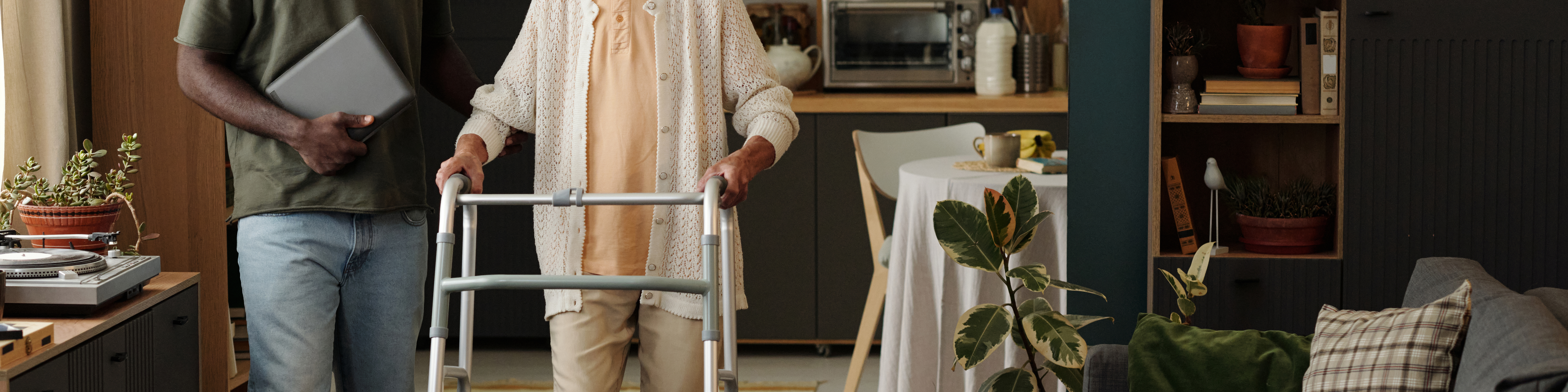 Senior Caucasian woman using walker for mobility support standing next to young adult Black man holding laptop in modern home kitchen setting, both partially visible