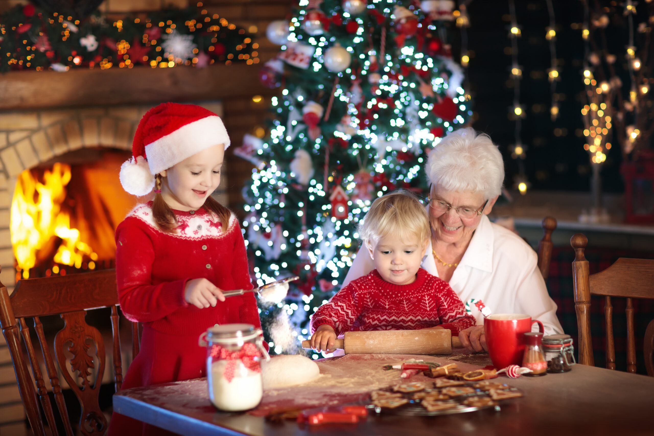 A young girl in a Santa hat and a boy in a red sweater are making Christmas cookies with their grandmother at a table. Behind them, a decorated Christmas tree and a lit fireplace create a festive atmosphere, with lights and ornaments enhancing the cozy setting.